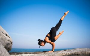 Donna che esegue una posizione yoga su una spiaggia rocciosa con vista sull'oceano e cielo azzurro limpido.