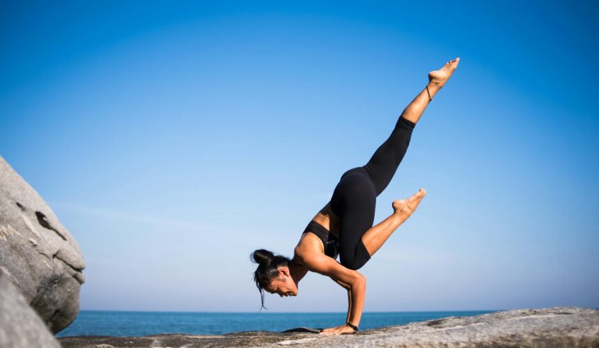 Mujer realizando una pose de yoga en una playa rocosa con vista al mar y cielo azul claro.