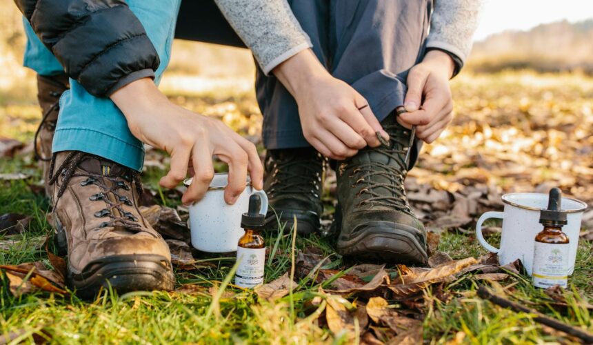 Primer plano de manos ajustando botas de montaña junto a aceite de CBD y tazas de café al aire libre.