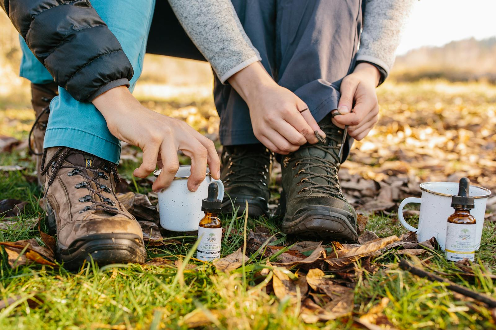 Close-up of hands adjusting hiking boots next to CBD oil and coffee cups outdoors.