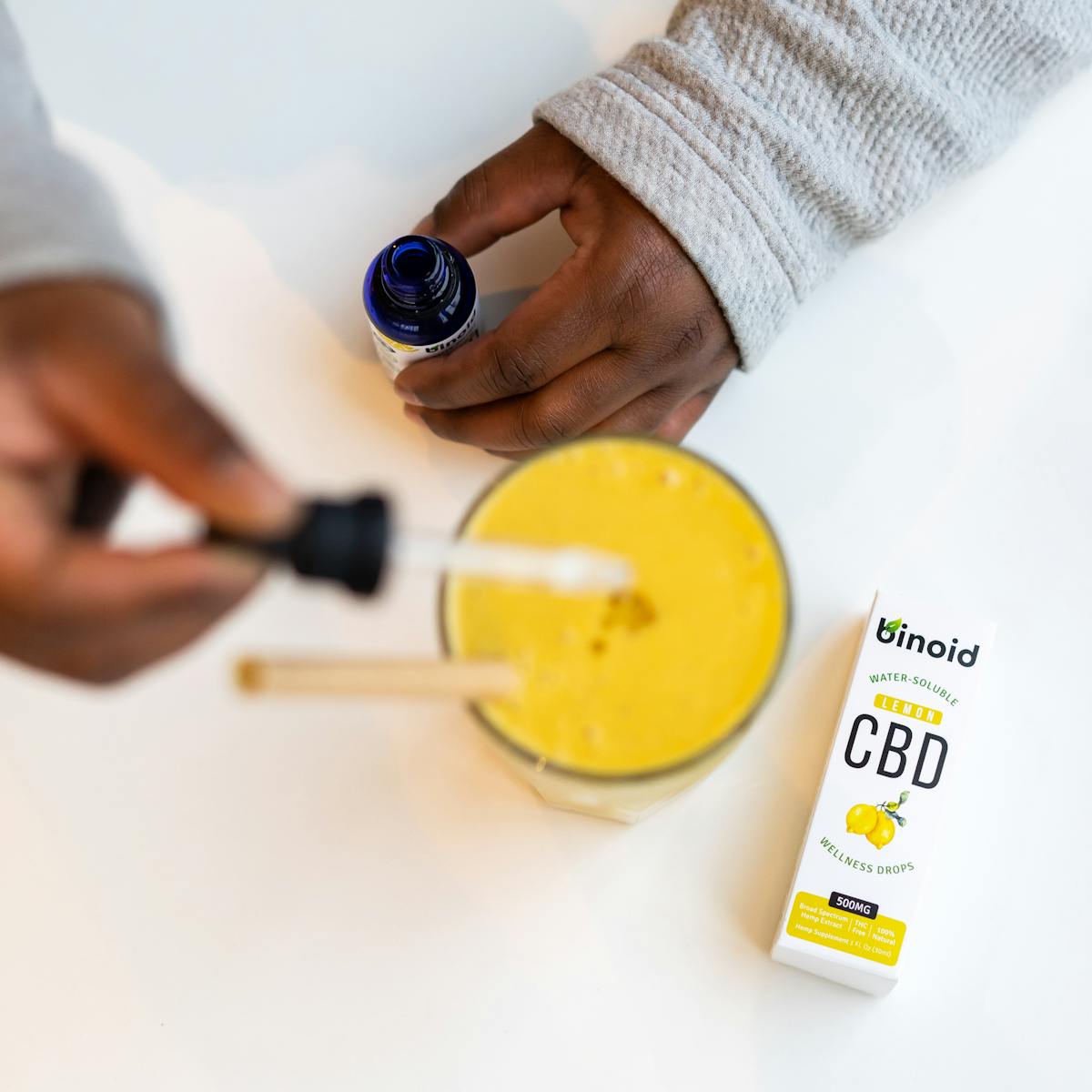 Close-up of hands using CBD oil with lemon smoothie, emphasizing wellness.