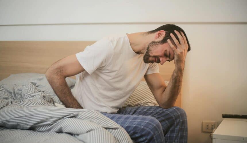 A young man in pajamas holding his head, sitting on a bed, appears to be experiencing a headache.