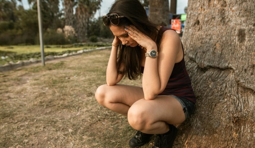 A woman kneels by a tree holding her head, expressing stress or anxiety in an outdoor park.